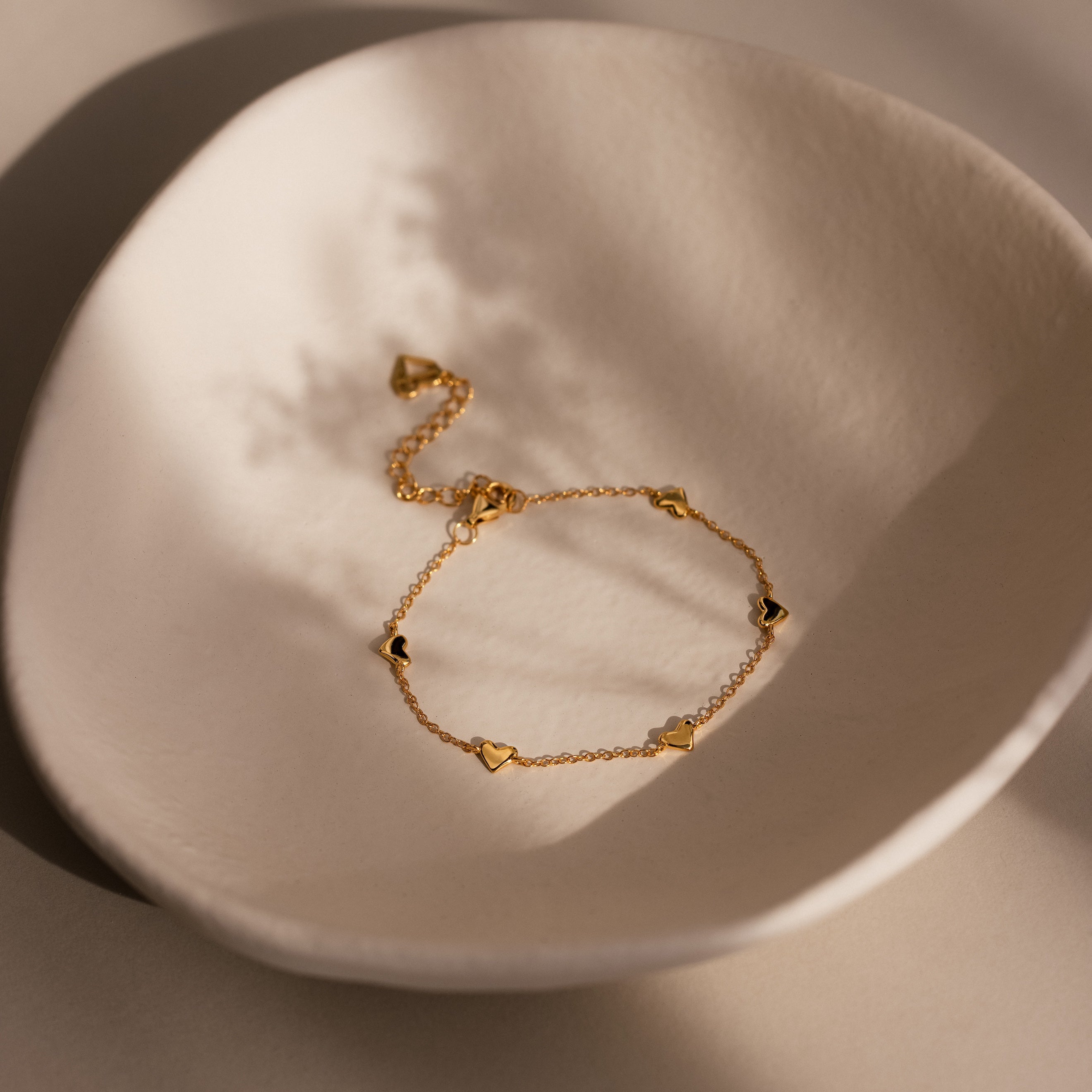 Heart Station Bracelet in 18K Gold, featuring delicate heart charms, is a romantic stacking piece shown on a textured off-white ceramic dish under soft light.