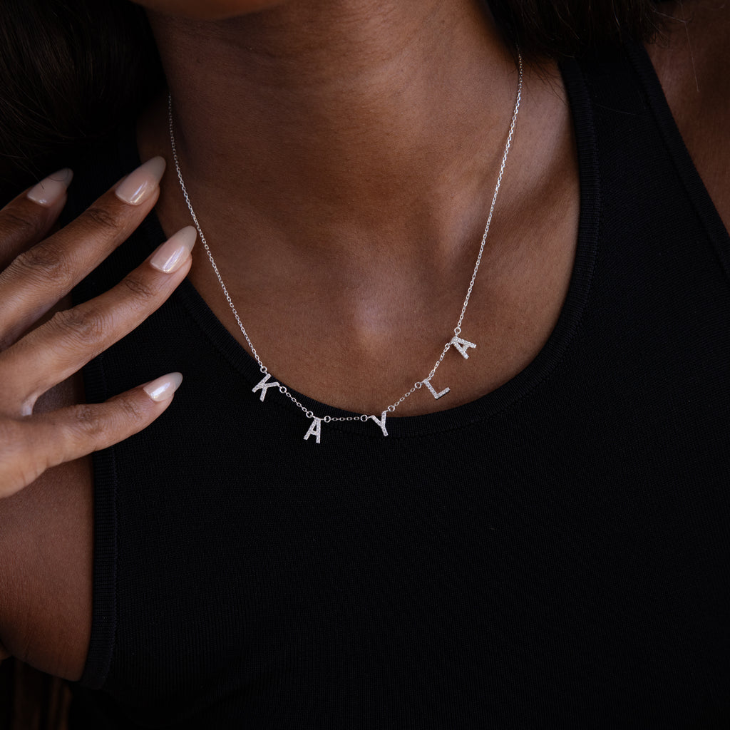 A woman wearing a Pave Letter Necklace in Sterling Silver with "KAYLA" charms and a black tank top, highlighting her love for delicate jewelry.