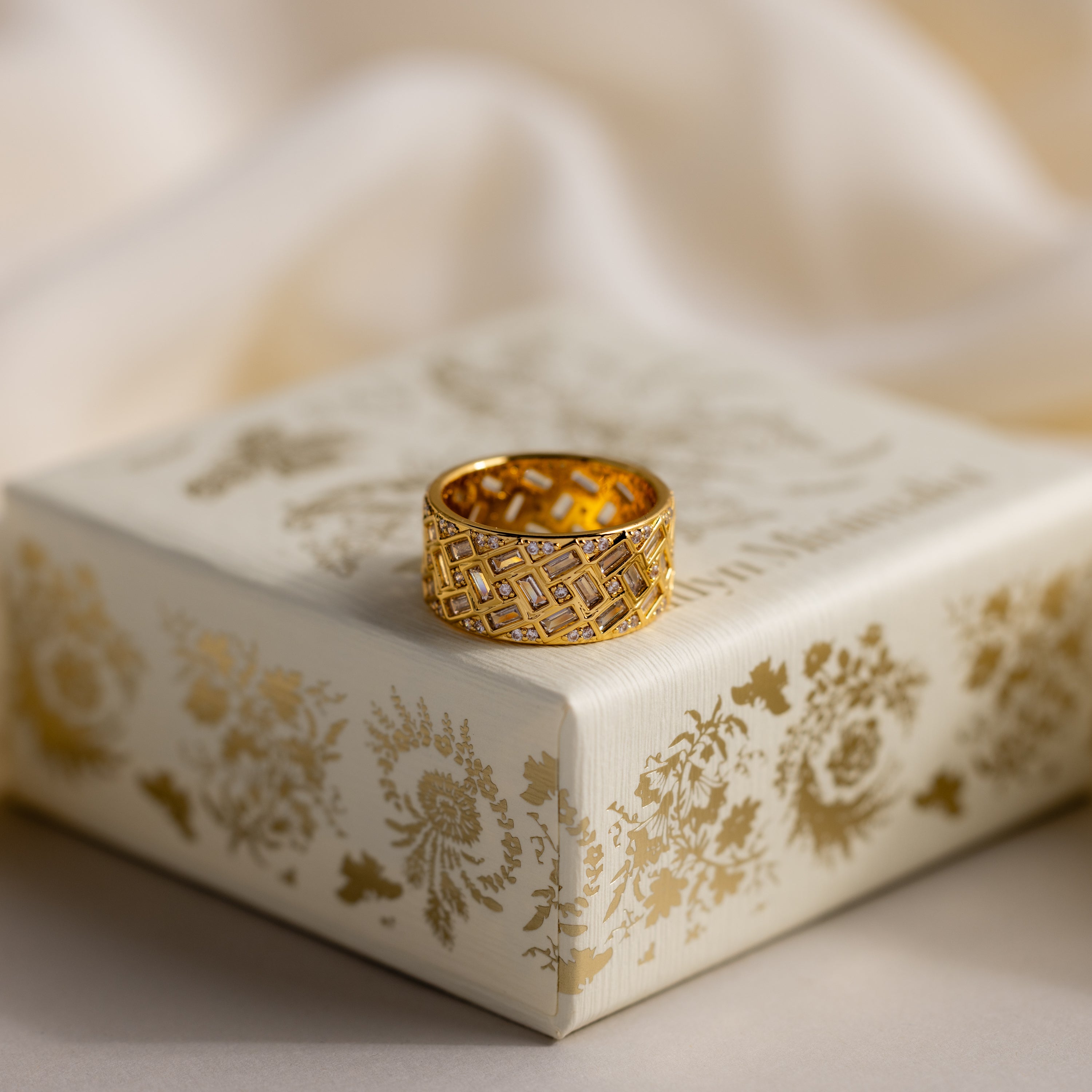 The Mosaic Diamond Baguette Ring, featuring an intricate geometric design, rests on a decorative cream and gold floral box.
