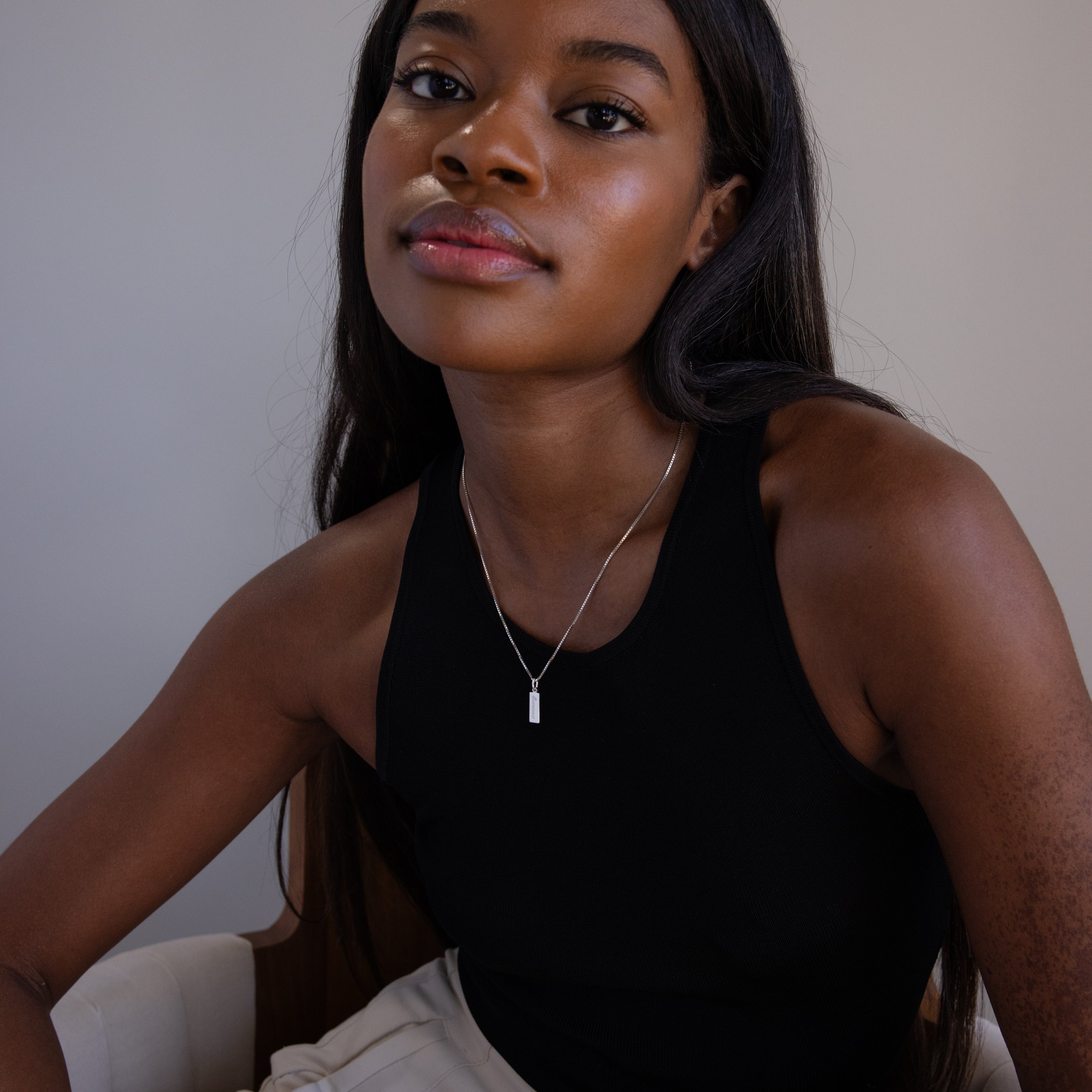 A woman with long hair sits on a white chair, wearing a black sleeveless top and the Dainty Tag Name Necklace.