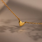 The Heart Station Necklace features a small, smooth gold heart pendant displayed on a soft beige background.