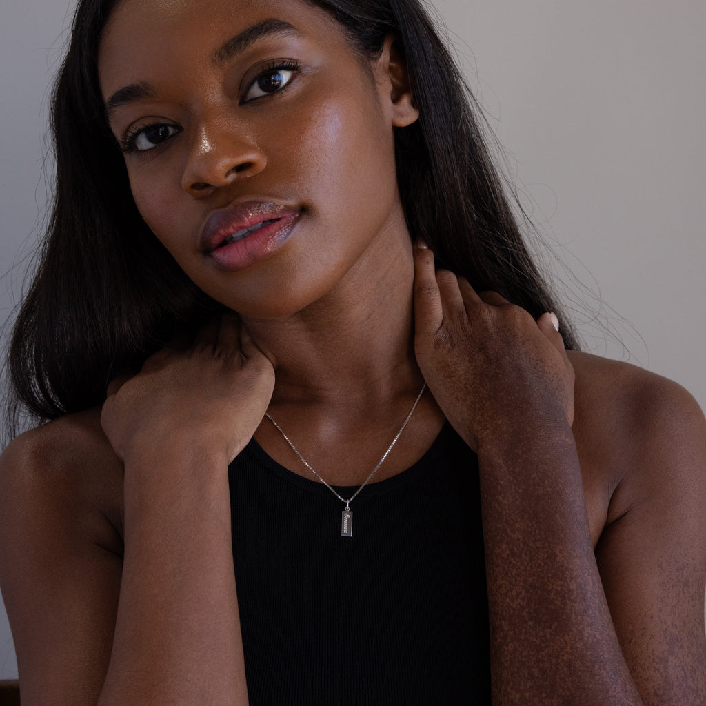 A woman in a black top touches her neck while wearing the Dainty Tag Name Necklace in Sterling Silver, featuring a rectangular pendant on delicate chains.