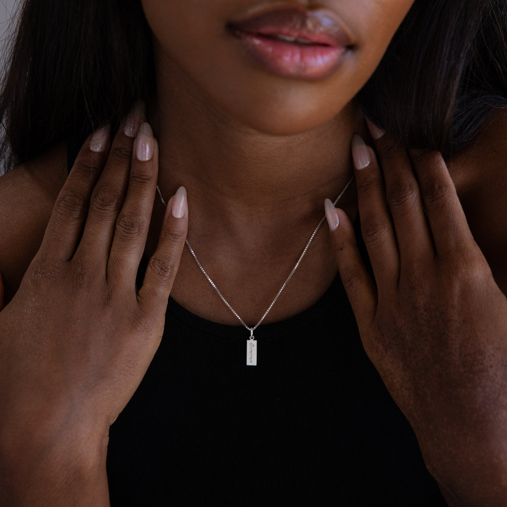 A woman wearing the Dainty Tag Name Necklace in Sterling Silver and a black top touches her collarbone with both hands. The delicate chains lend a touch of elegance to her look.