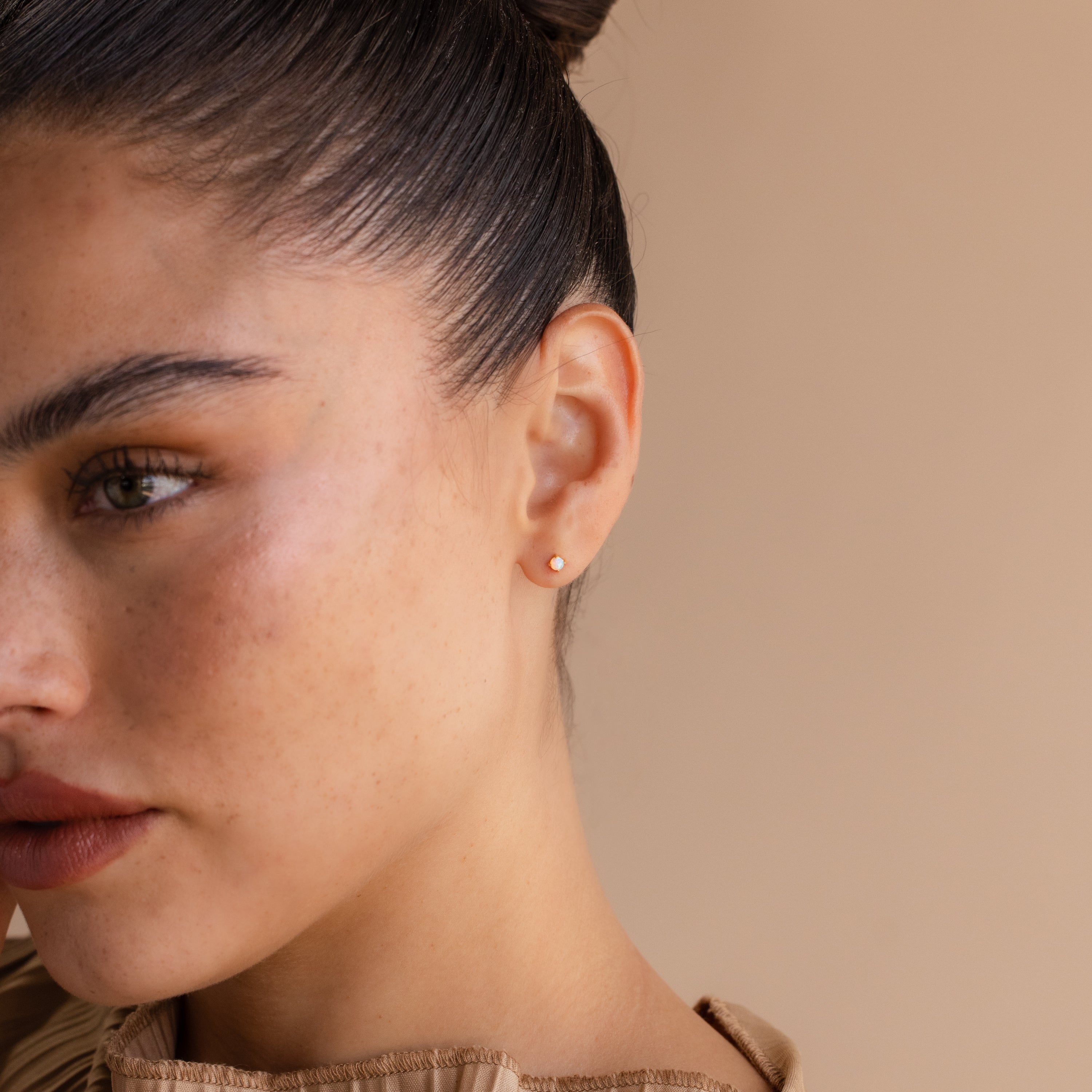 Close-up of a woman with a smooth complexion and hair in a bun, wearing Opal Flat Back Studs in 18K Gold against a beige background.