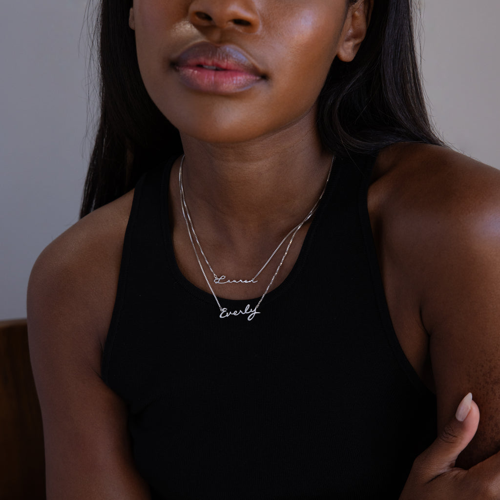 Woman in black sleeveless top wears two silver Callie Box Chain Name Necklaces, featuring “Ernie” and “Everly” nameplates on box chains. This customizable necklace is perfect for layering and adding a personal touch to any look.