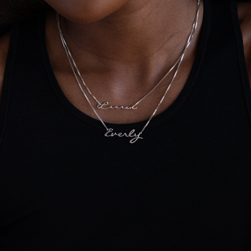 A person wearing two Callie Box Chain Name Necklaces in Sterling Silver—one customized with "Laurel" and the other with "Everly"—layered over a black tank top.
