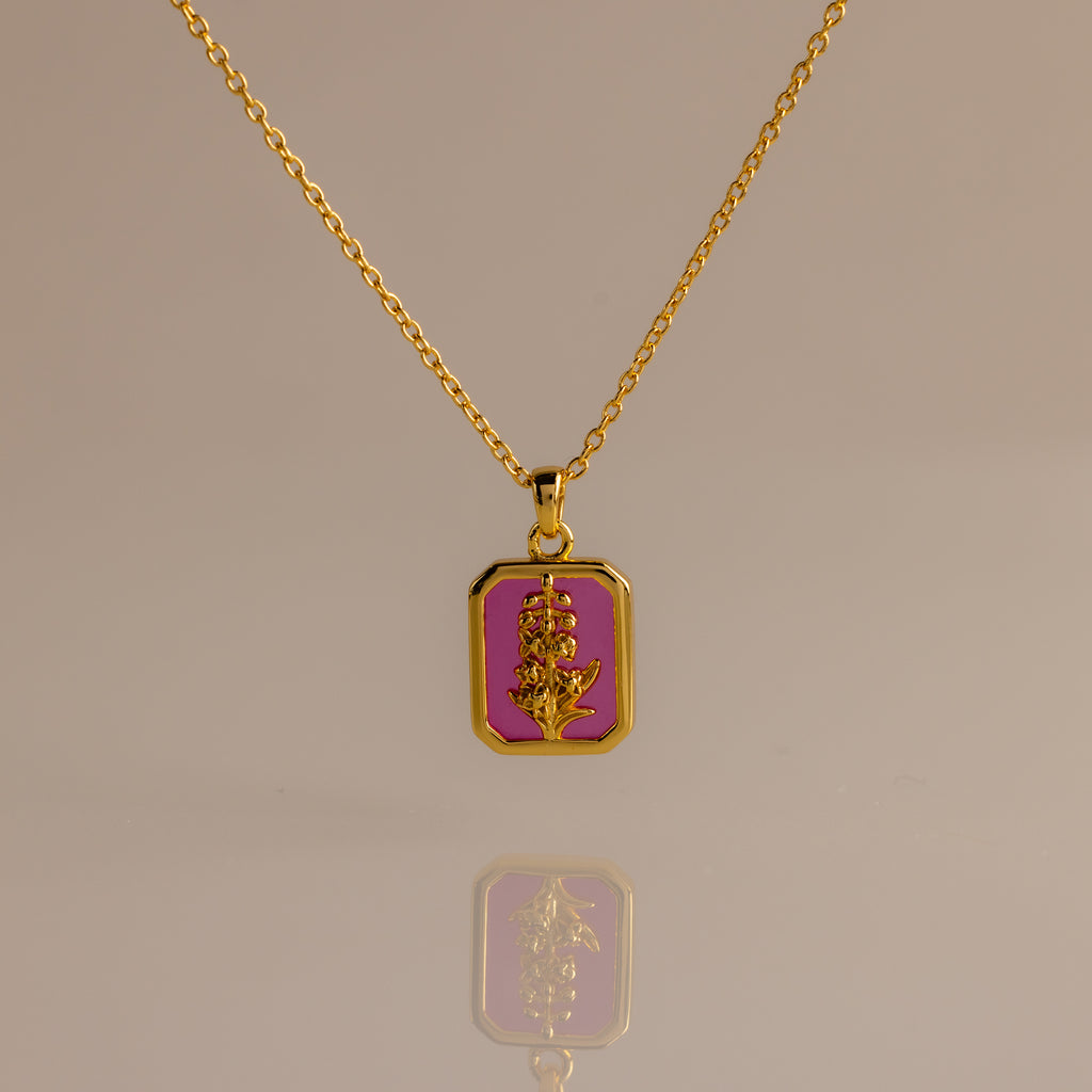 The Ruby Birth Flower Charm Necklace, a gold chain with a rectangular pink pendant featuring a gold floral design, is elegantly displayed on a neutral background.