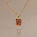 The Ruby Birth Flower Charm Necklace, a gold chain with a rectangular pink pendant featuring a gold floral design, is elegantly displayed on a neutral background.