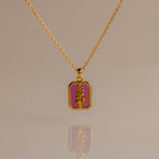 The Ruby Birth Flower Charm Necklace, a gold chain with a rectangular pink pendant featuring a gold floral design, is elegantly displayed on a neutral background.