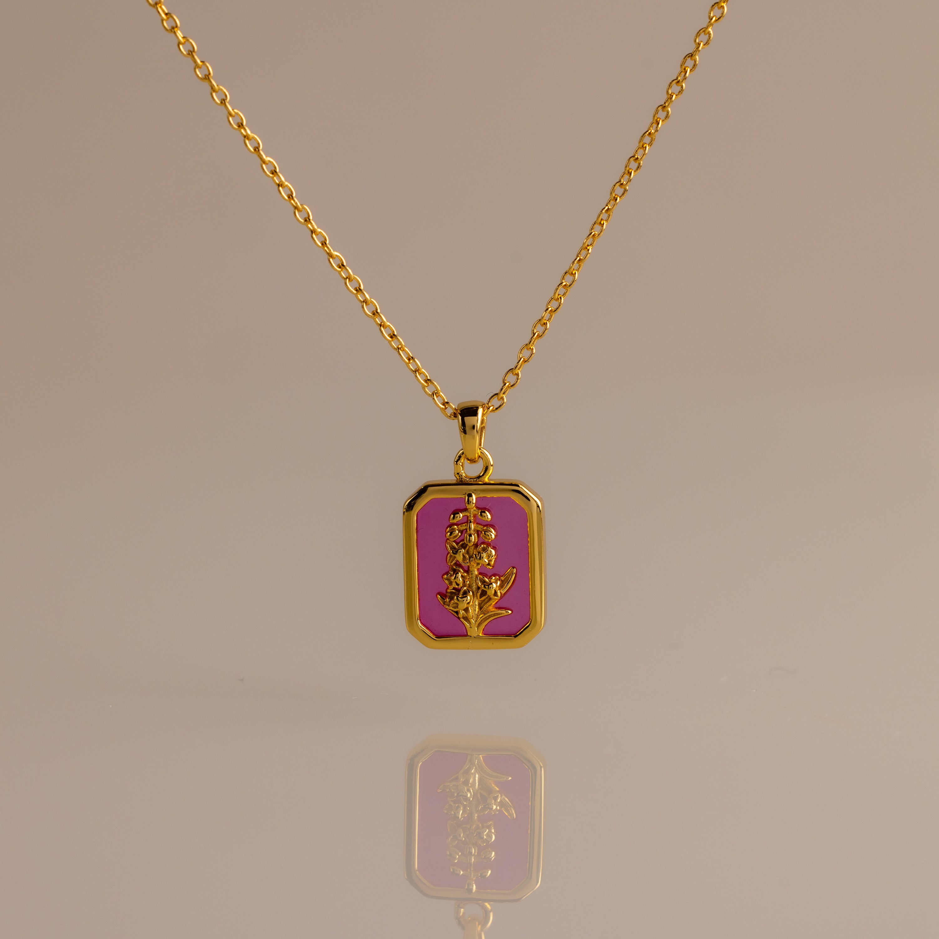 The Ruby Birth Flower Charm Necklace, a gold chain with a rectangular pink pendant featuring a gold floral design, is elegantly displayed on a neutral background.