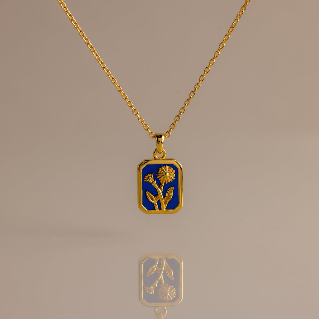 The Sapphire Birth Flower Charm Necklace features a sapphire-blue rectangular pendant with a delicate gold flower design, displayed on a neutral background.