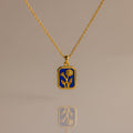 The Sapphire Birth Flower Charm Necklace features a sapphire-blue rectangular pendant with a delicate gold flower design, displayed on a neutral background.