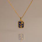 The Sapphire Birth Flower Charm Necklace features a sapphire-blue rectangular pendant with a delicate gold flower design, displayed on a neutral background.