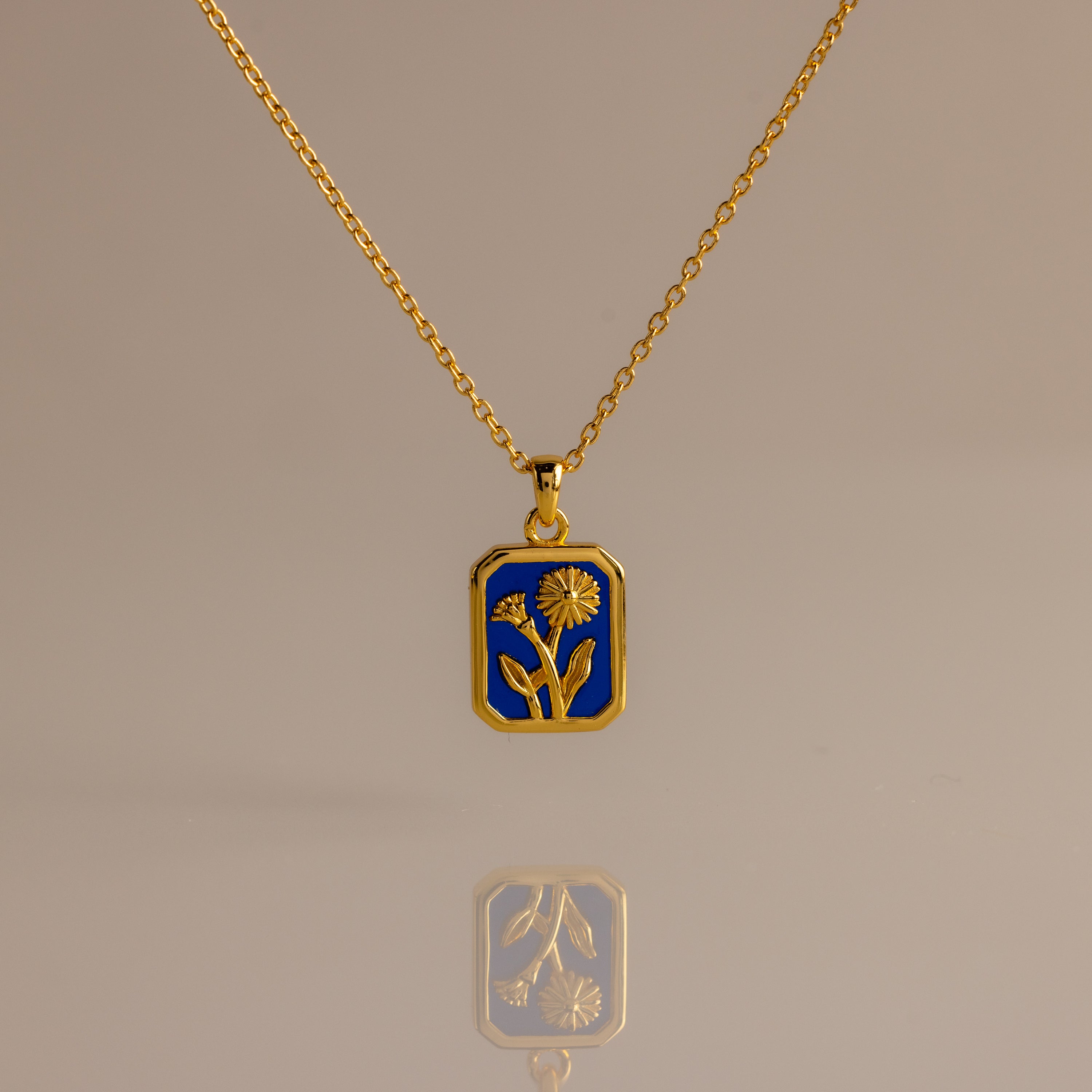 The Sapphire Birth Flower Charm Necklace features a sapphire-blue rectangular pendant with a delicate gold flower design, displayed on a neutral background.