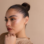 A woman with brown hair in a bun wears Opal Flat Back Studs in 18K Gold and a beige top, gazing thoughtfully to the side against a matching beige background.