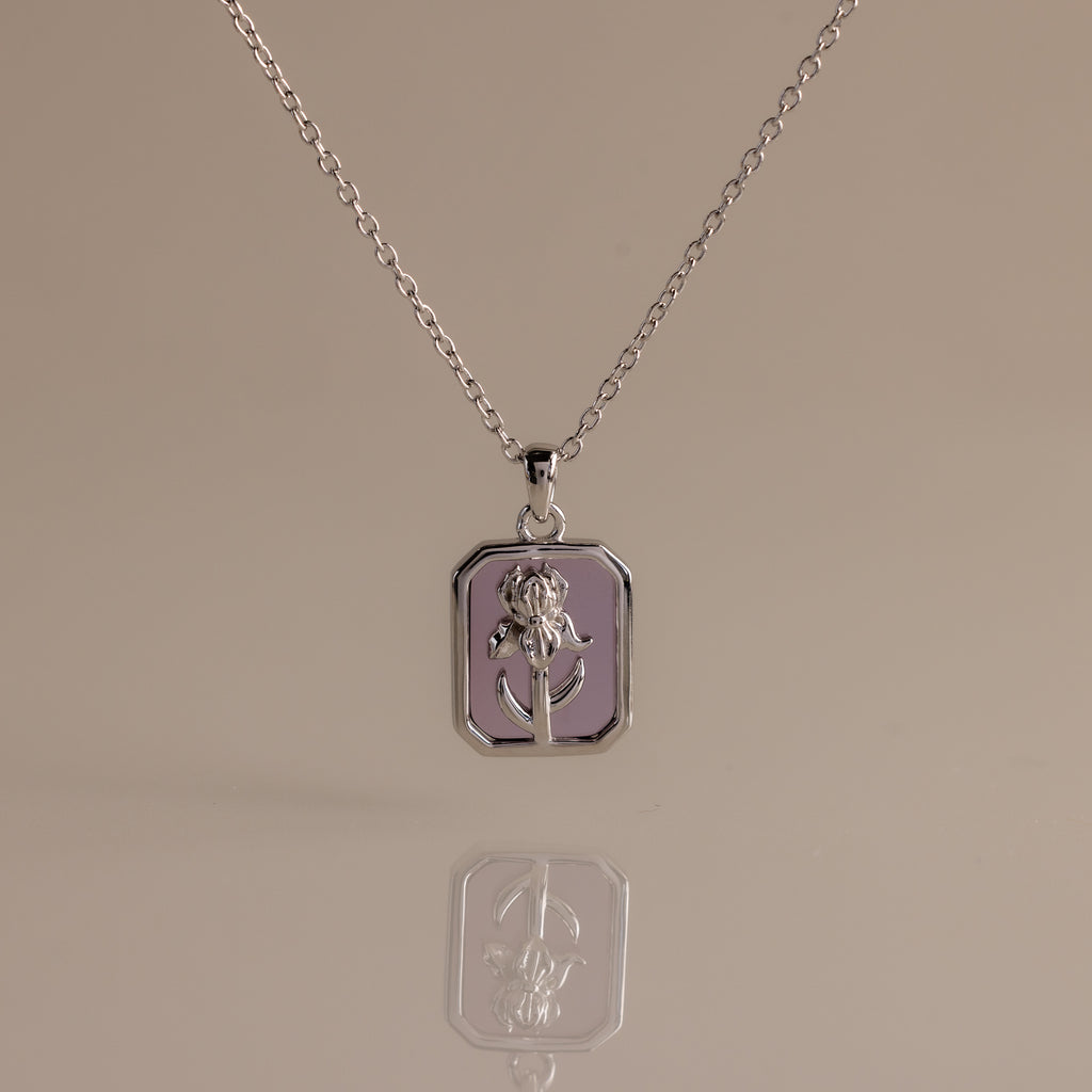 The Amethyst Birth Flower Charm Necklace features a silver rectangular pendant with a raised floral design, making it a perfect delicate jewelry piece. Displayed on a neutral background for an elegant presentation.