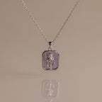 The Amethyst Birth Flower Charm Necklace features a silver rectangular pendant with a raised floral design, making it a perfect delicate jewelry piece. Displayed on a neutral background for an elegant presentation.