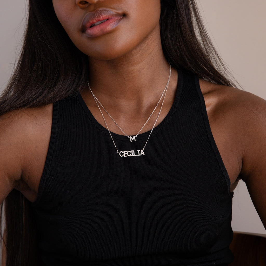 A woman in a black tank top wears two silver necklaces: a Pave Initial Necklace in Sterling Silver with an M and another spelling CECILIA.