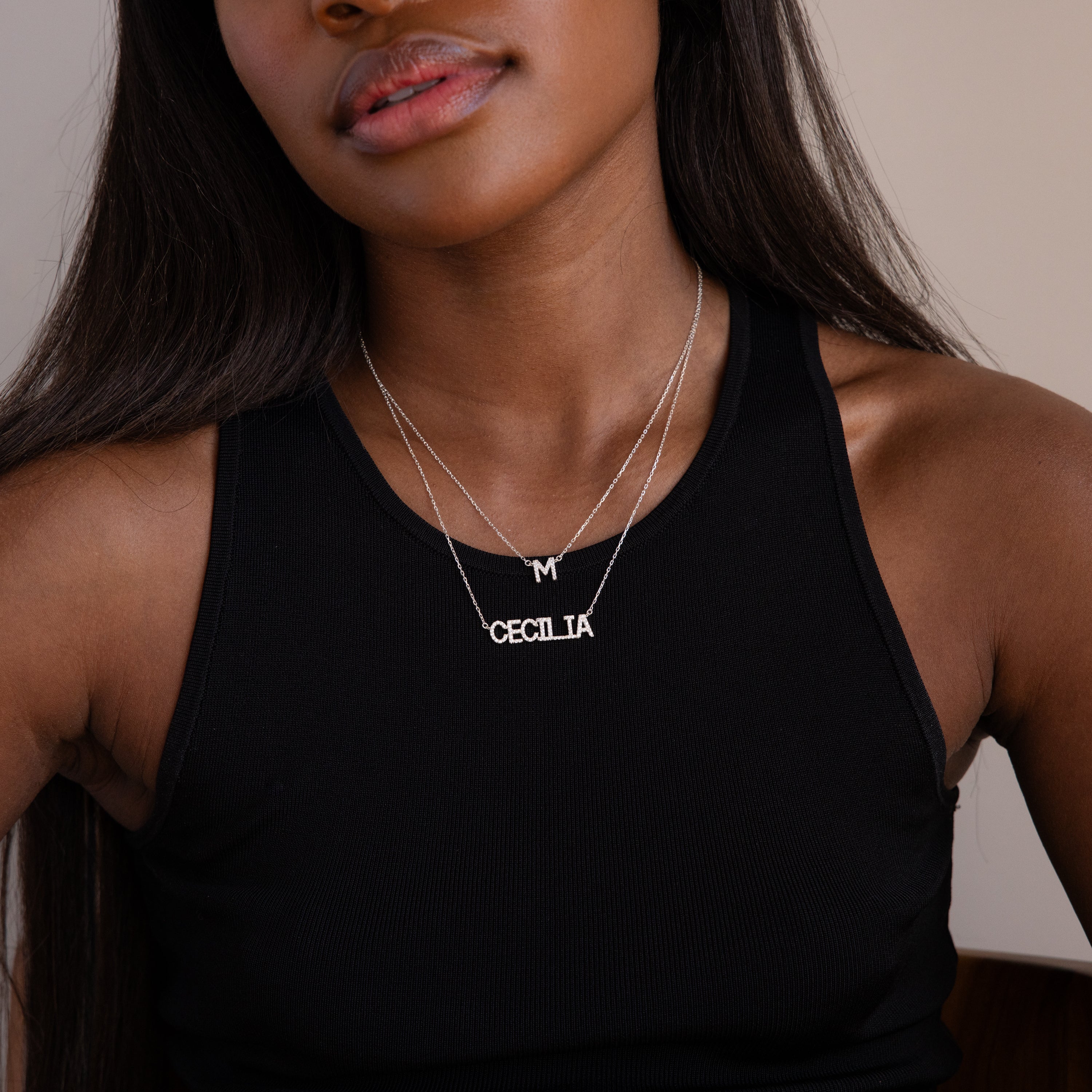 A woman in a black tank top wears two silver necklaces: a Pave Initial Necklace in Sterling Silver with an M and another spelling CECILIA.