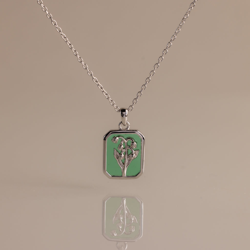 Silver necklace with a rectangular green emerald birth flower charm displayed on a reflective surface—I'm sorry, but there is no product description provided for further details.