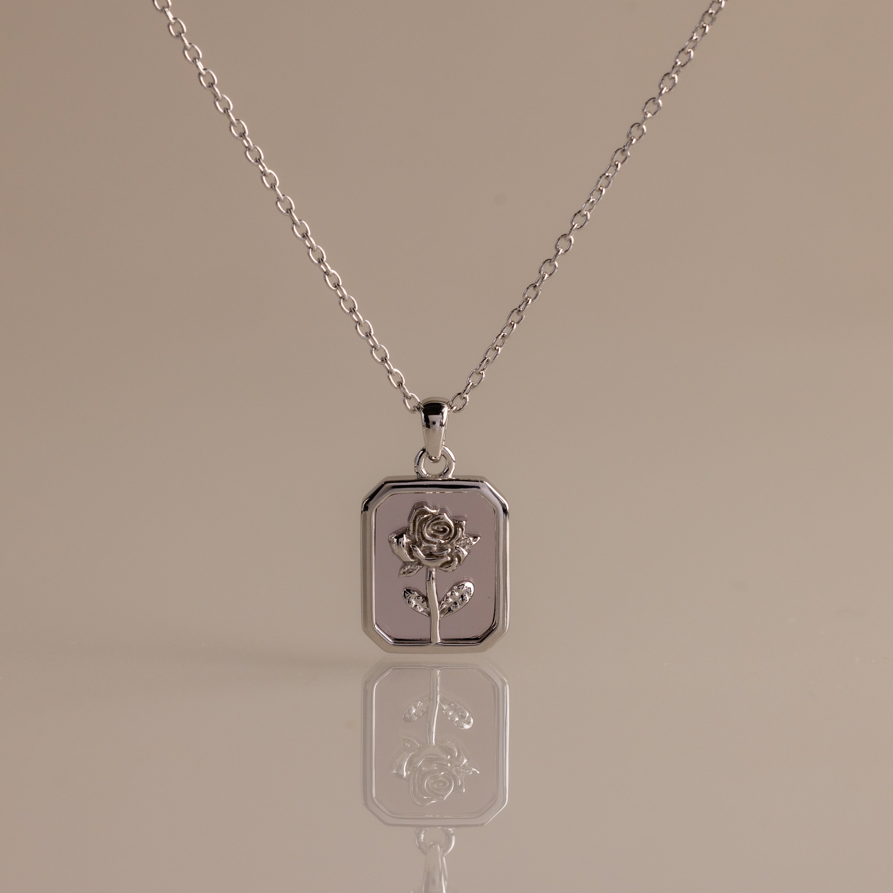 The Alexandrite Birth Flower Charm Necklace features a rectangular pendant with an engraved rose design, offering elegant style. Beautifully displayed on a neutral background, it’s the perfect accessory for a refined look.