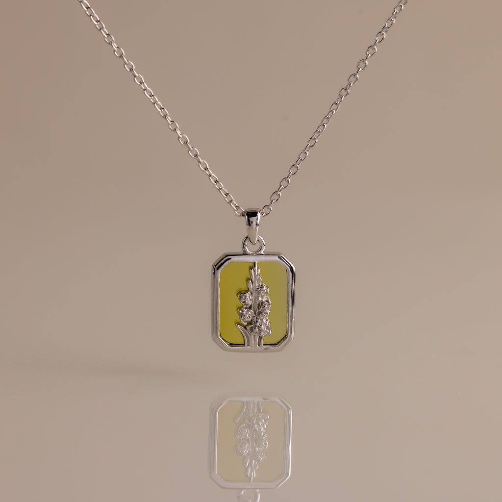 The Peridot Birth Flower Charm Necklace features a rectangular floral pendant on a yellow background, making it an ideal gift for nature lovers and flower jewelry enthusiasts. Displayed on a plain surface.