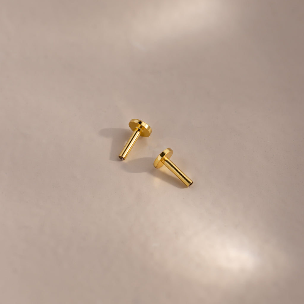 Two 18k gold Flat Back Posts lying on a matte surface