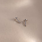 Two silver flatback earring posts resting on a soft beige surface with gentle lighting and shadows.