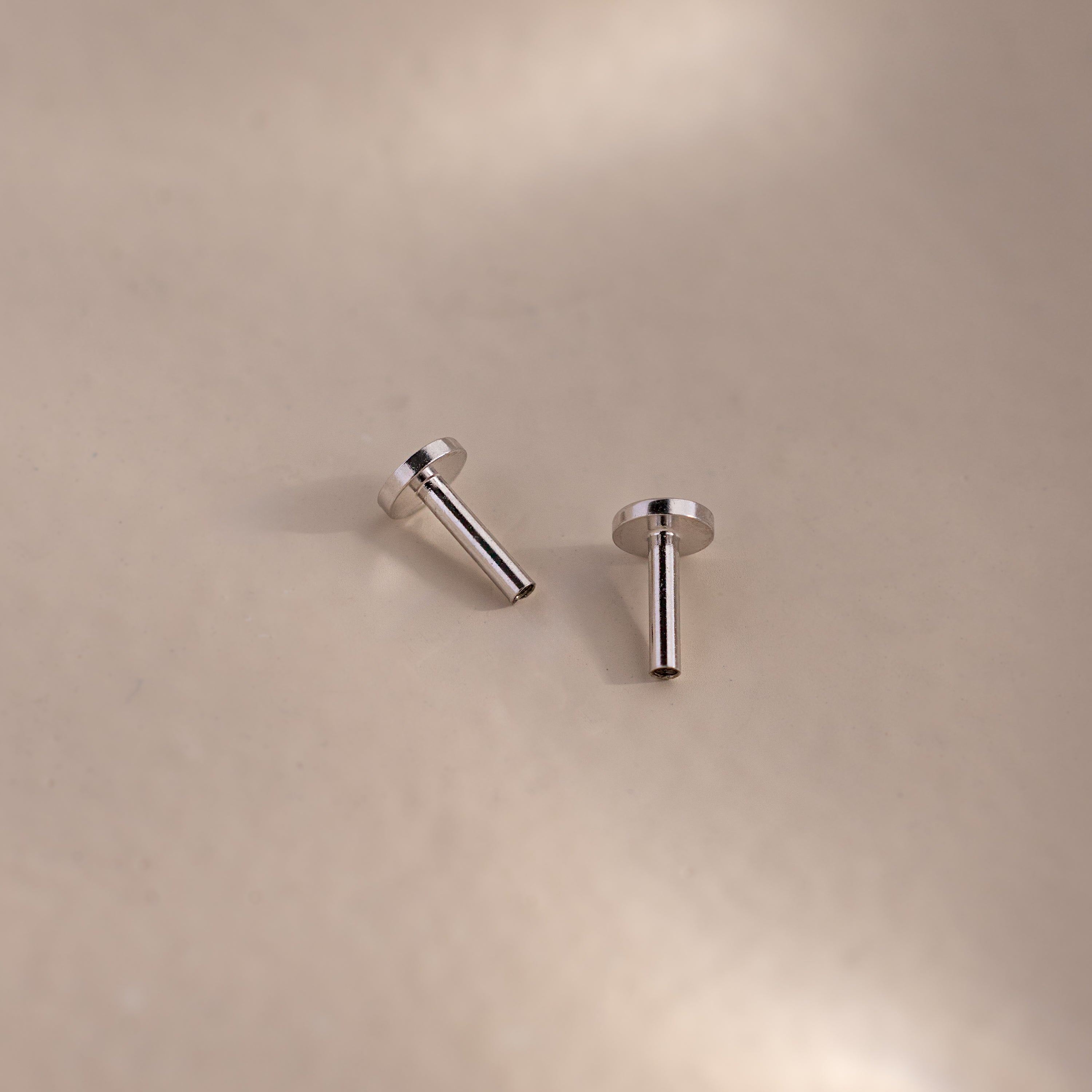 Two silver flatback earring posts resting on a soft beige surface with gentle lighting and shadows.