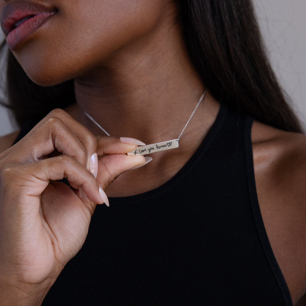 A woman wears a black sleeveless top and holds the Handwriting Bar Necklace in Sterling Silver engraved with "Love you forever"—an elegant, personalized jewelry piece ideal for a heartfelt gift.