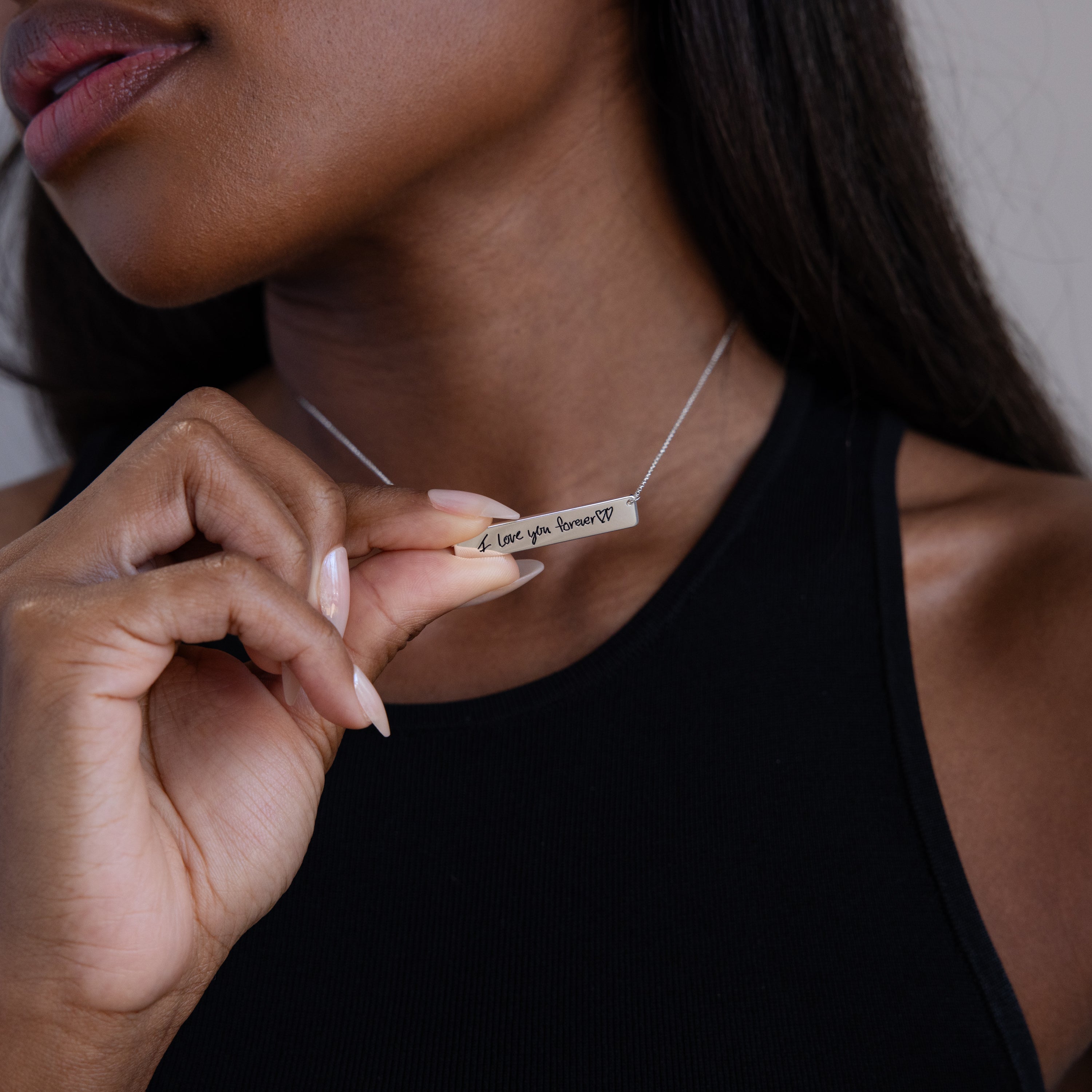 A woman wears a black sleeveless top and holds the Handwriting Bar Necklace in Sterling Silver engraved with "Love you forever"—an elegant, personalized jewelry piece ideal for a heartfelt gift.