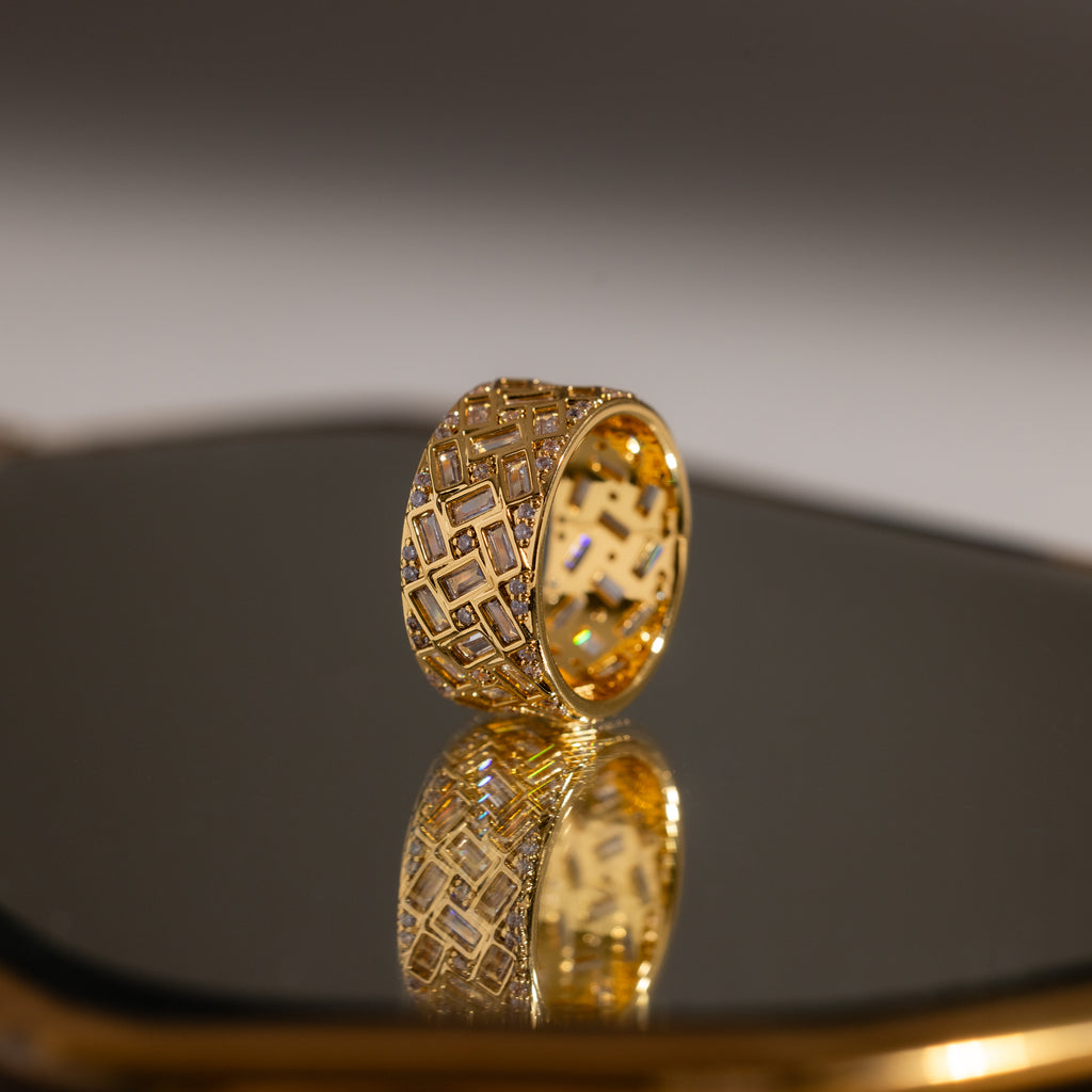 The Mosaic Diamond Baguette Ring features a bold, geometric cutout design in gold, beautifully displayed as its wide band elegantly reflects on a shiny surface.
