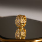 The Mosaic Diamond Baguette Ring features a bold, geometric cutout design in gold, beautifully displayed as its wide band elegantly reflects on a shiny surface.