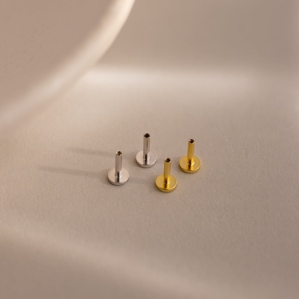 Four internally threaded flatback earring posts in silver and gold finishes, arranged on a neutral-toned surface with soft shadows.