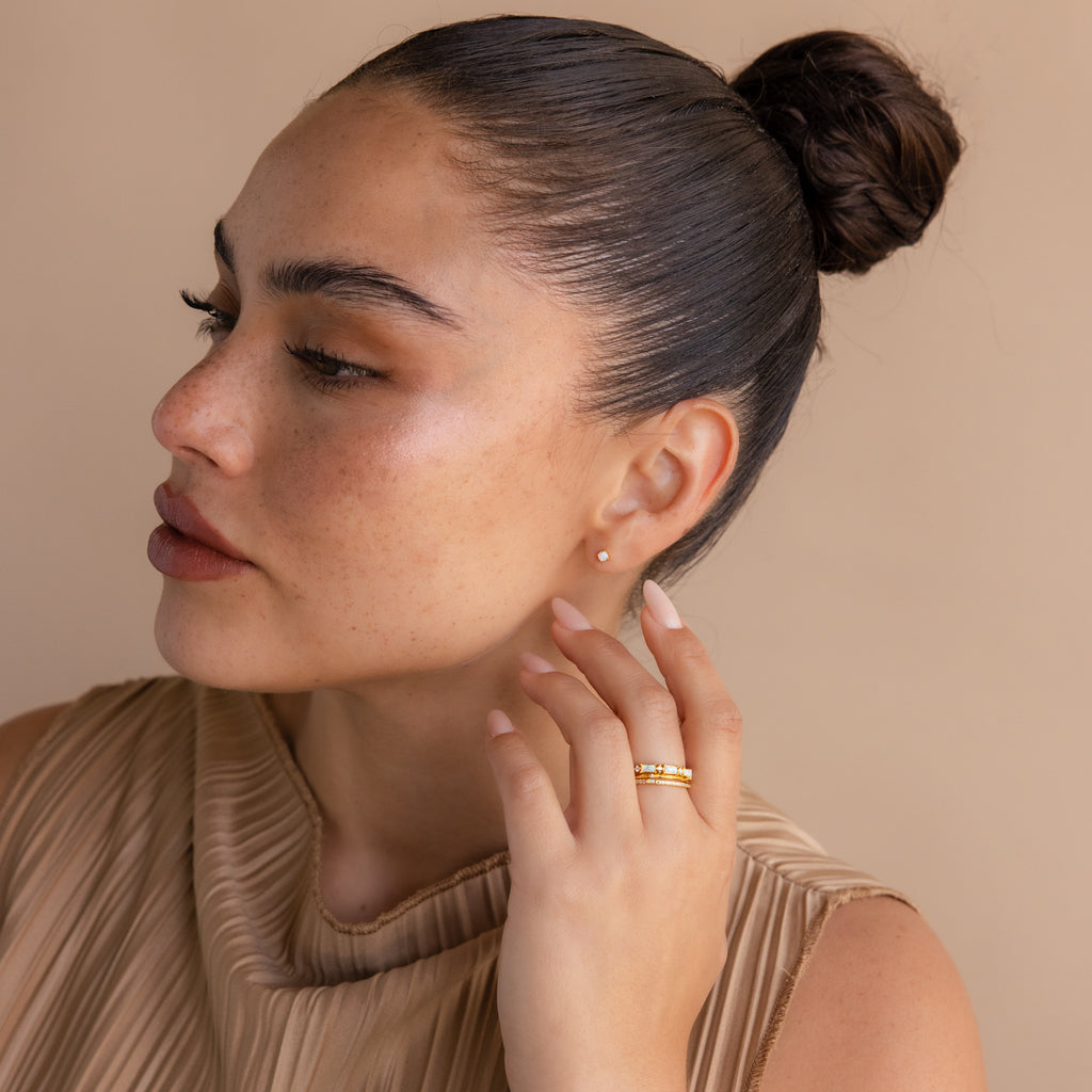 Woman with dark slicked-back hair in a bun, wearing a beige top, Opal Flat Back Studs in 18K Gold, and gold rings, looking to the side.