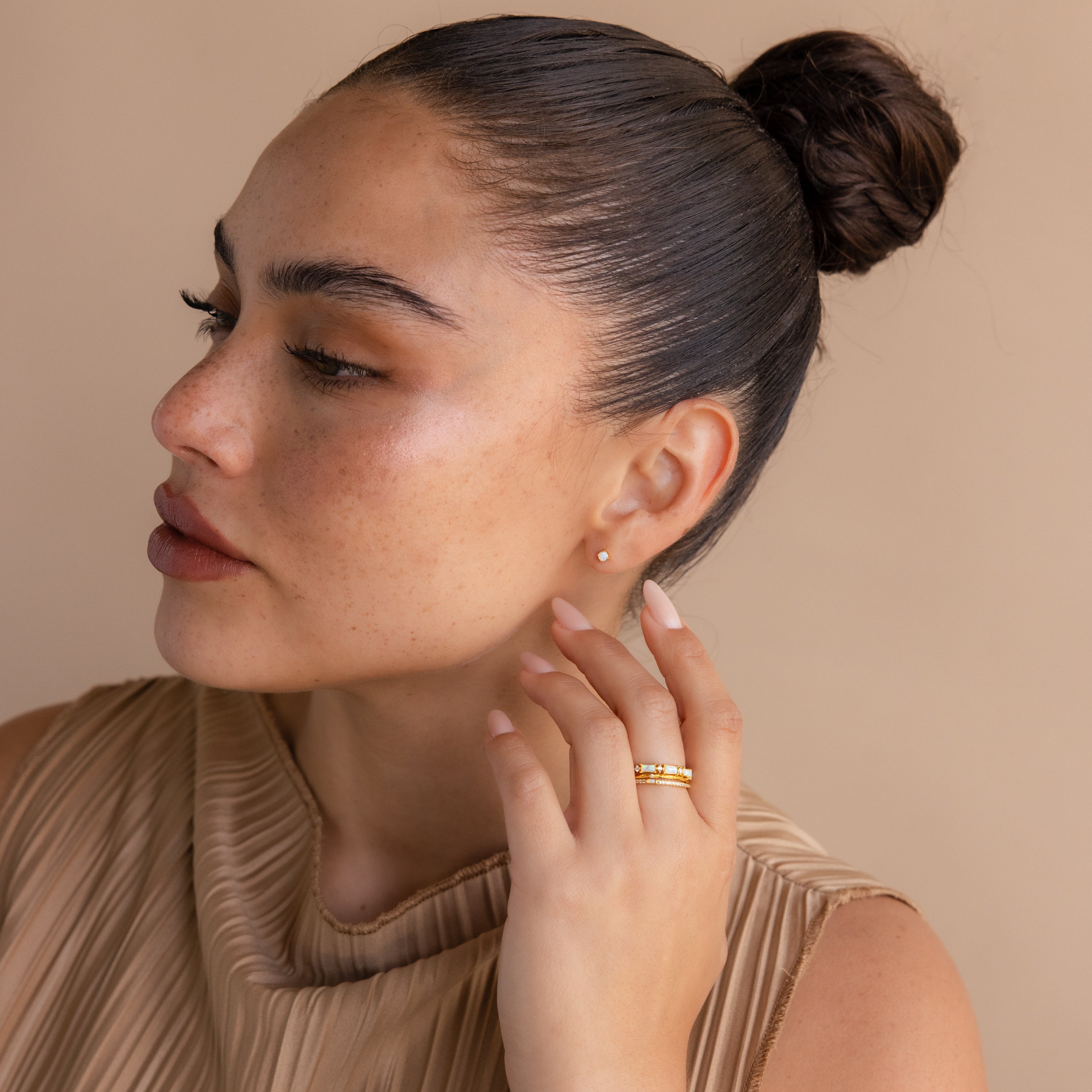 Woman with dark slicked-back hair in a bun, wearing a beige top, Opal Flat Back Studs in 18K Gold, and gold rings, looking to the side.