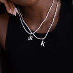 A person wears the Newsletter Pearl Initial Necklace in Sterling Silver with customizable R and K charms over a black top, showcasing elegant minimalist jewelry.