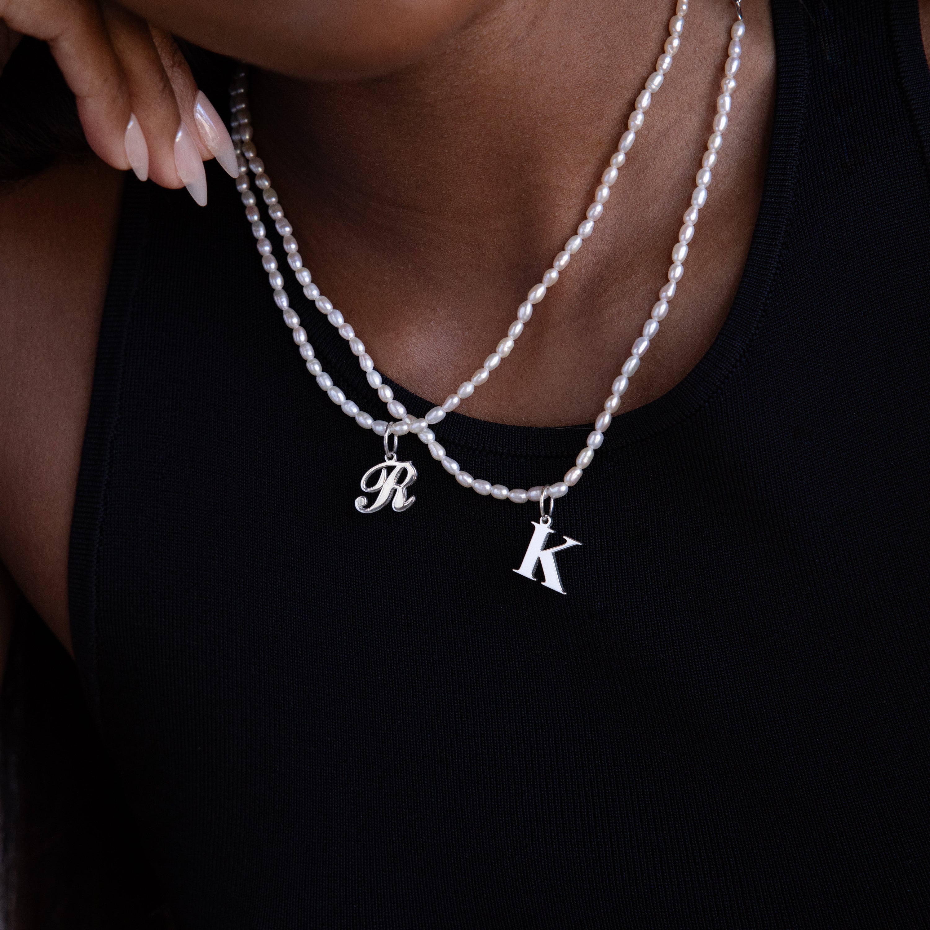 A person wears the Newsletter Pearl Initial Necklace in Sterling Silver with customizable R and K charms over a black top, showcasing elegant minimalist jewelry.