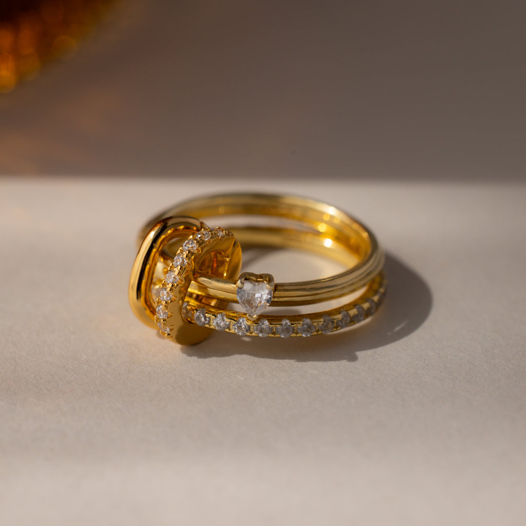 The Multipurpose Pave Ring Lock Set in 18K Gold features three gold rings with diamonds and a heart-shaped gemstone, finished with a pavé-detailed clasp, stacked elegantly on a light surface.