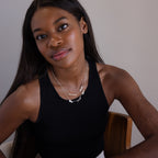 A woman with long hair in a black tank top sits facing the camera, wearing layered silver necklaces including the Carrie Multiple Name Necklace in Sterling Silver.