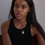 Woman with long hair in a black tank top wears the Oval Flower Necklace in Sterling Silver, sitting and looking at the camera.