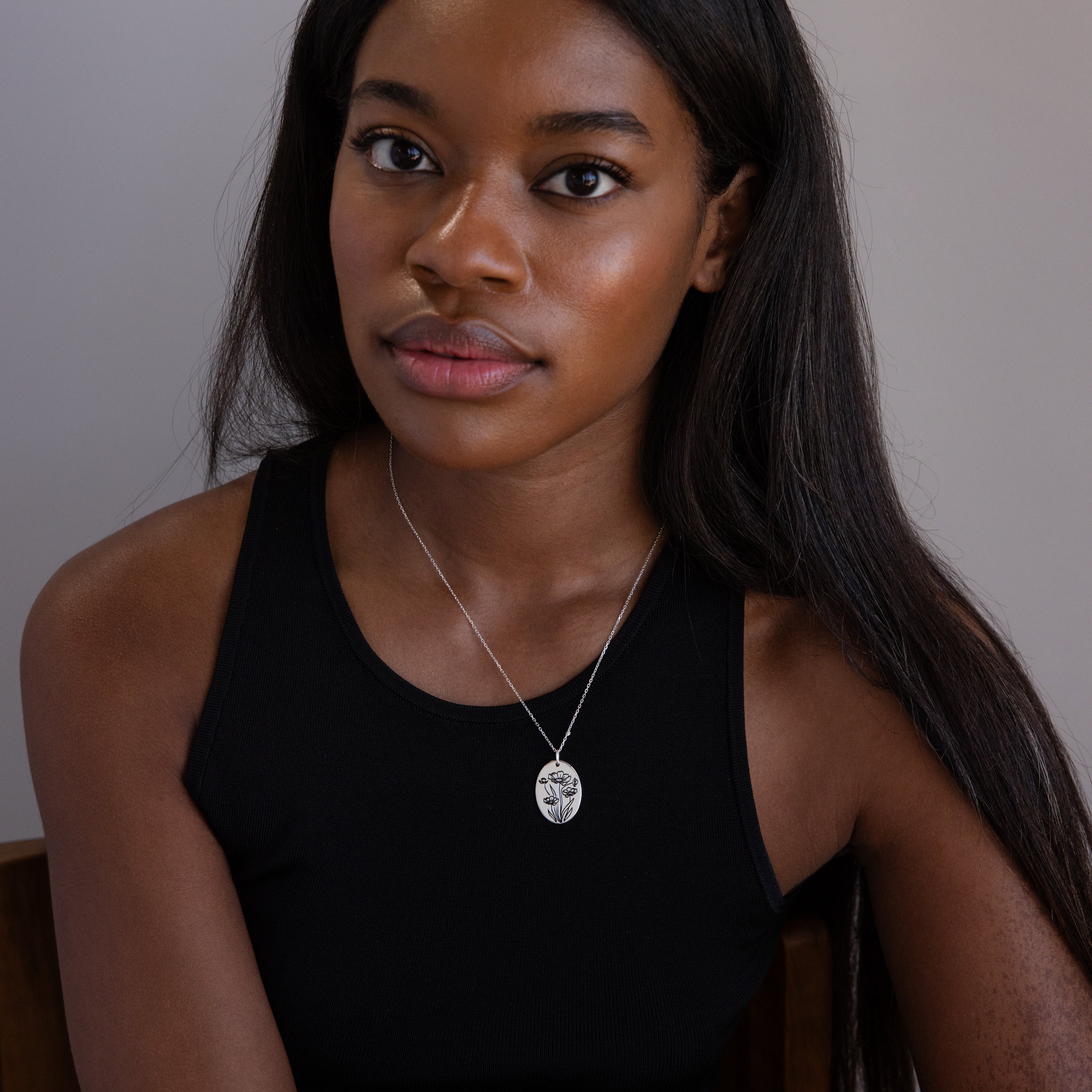 Woman with long hair in a black tank top wears the Oval Flower Necklace in Sterling Silver, sitting and looking at the camera.