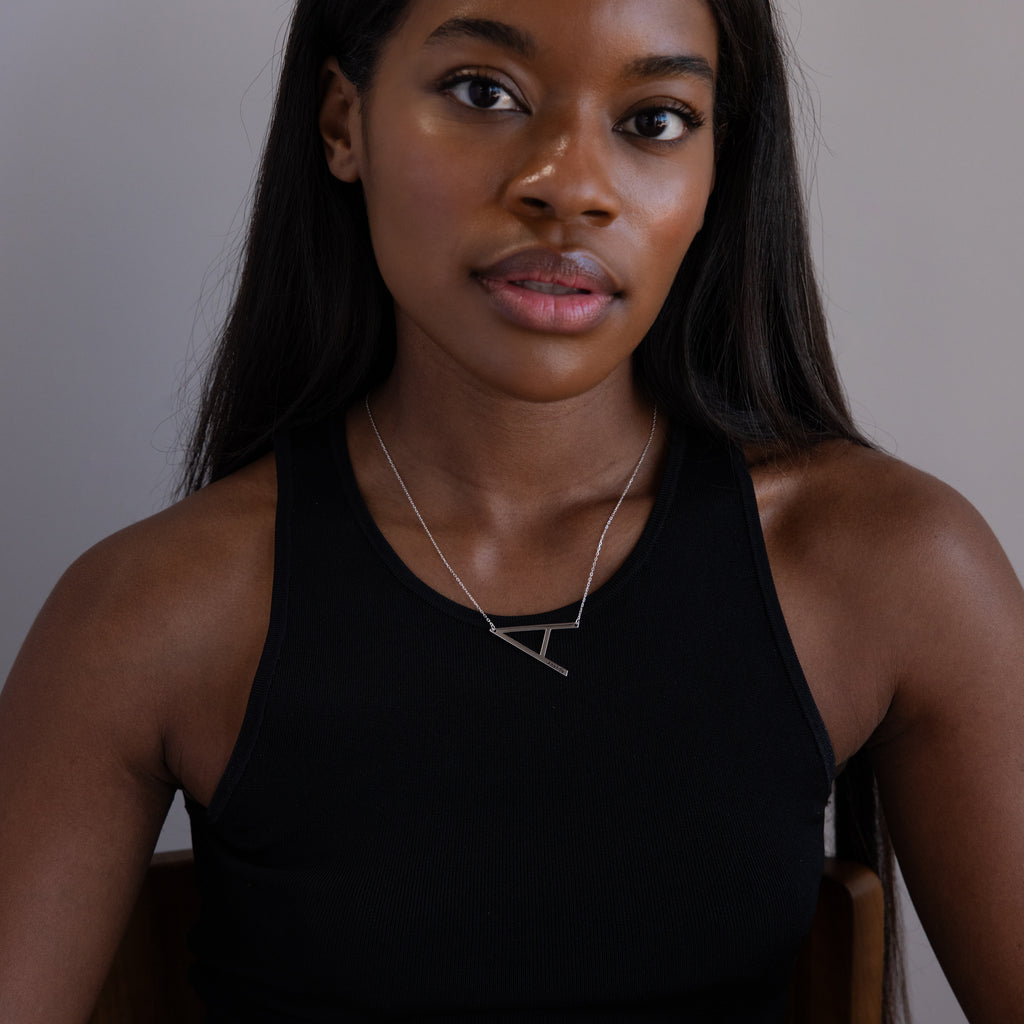 Woman in a black sleeveless top wearing the Large Sideways Initial Necklace in Sterling Silver, sitting and looking at the camera.