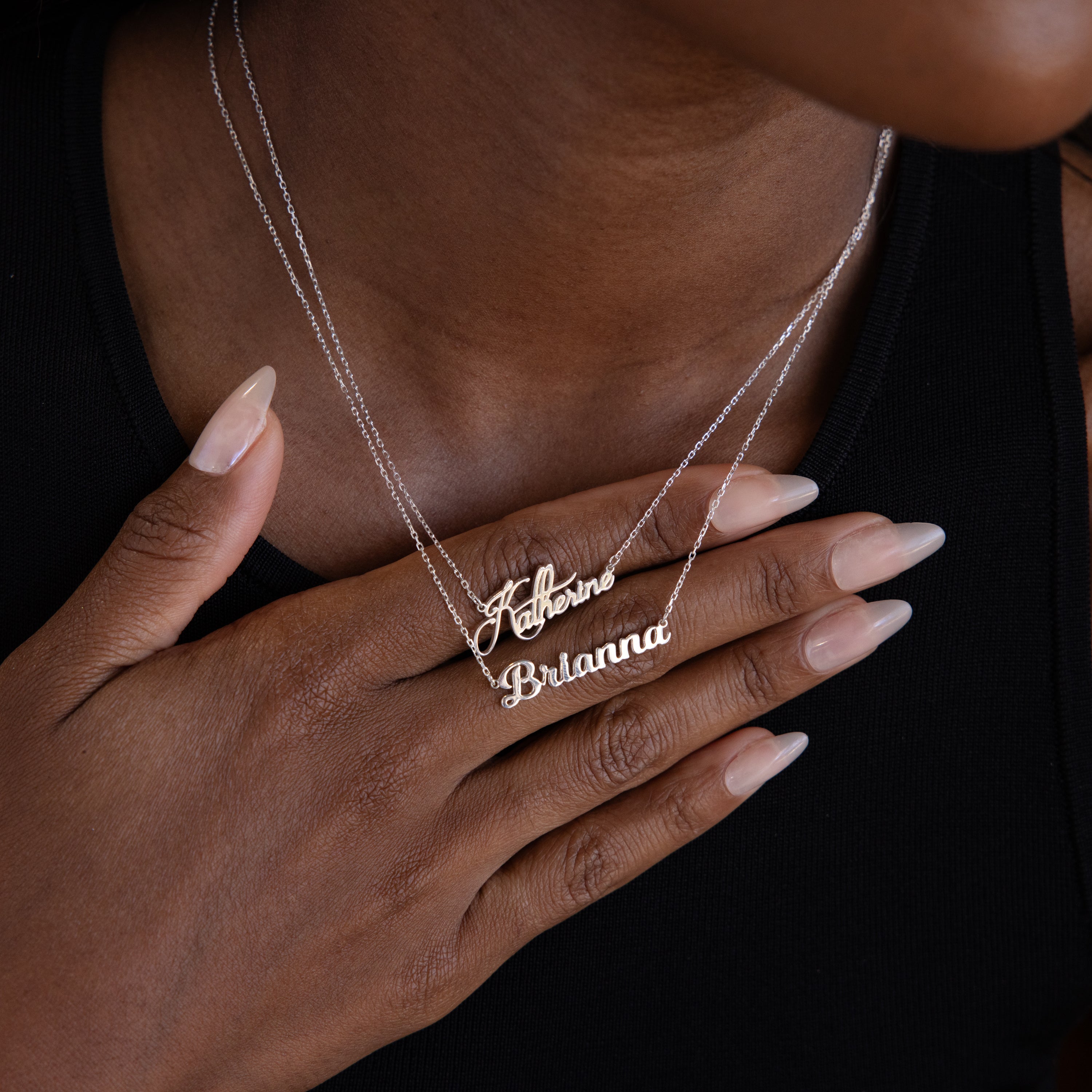 A person wearing two silver necklaces—a Mellow Name Necklace in Sterling Silver reading Katherine and another name necklace for Brianna—with a hand on their chest.