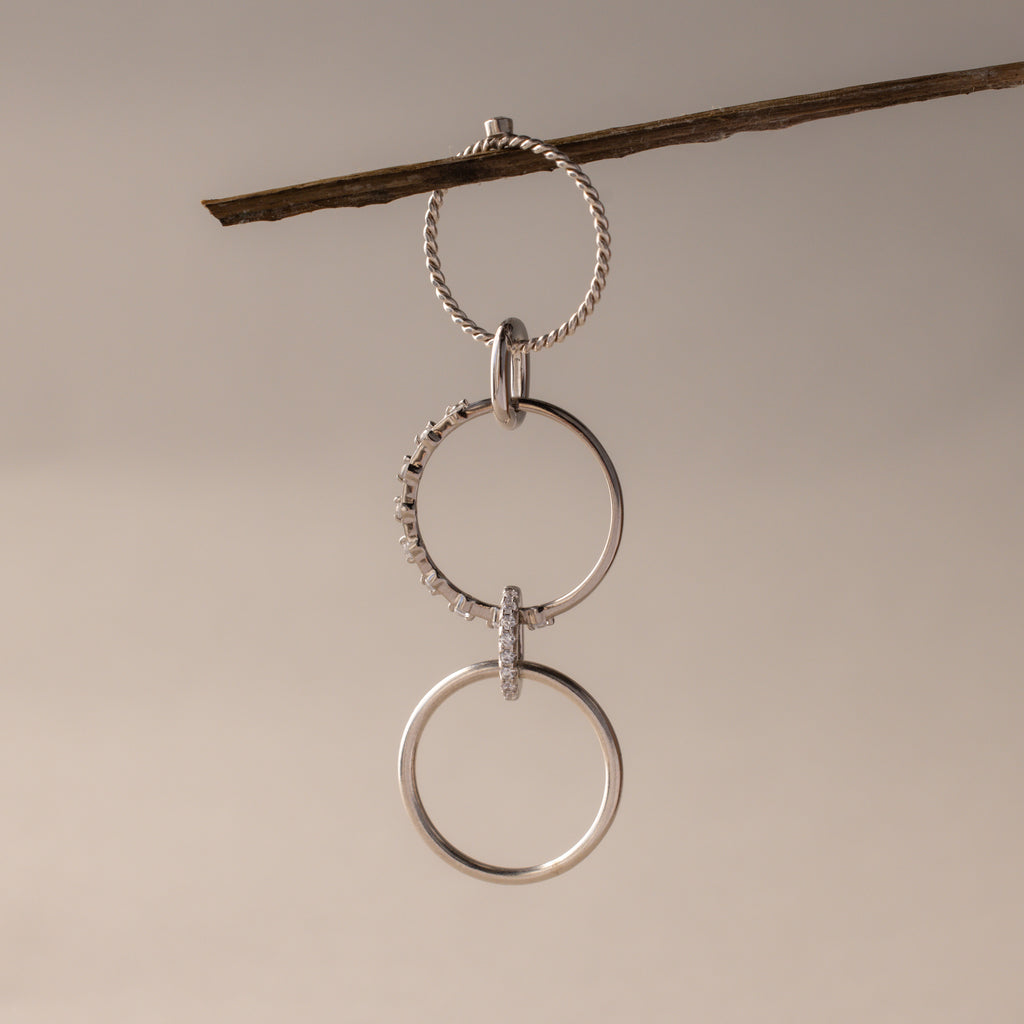Three silver rings, including the Multipurpose Pave Ring Lock Set in White Gold, hang linked from a twig against a neutral background—highlighting versatile jewelry customization.