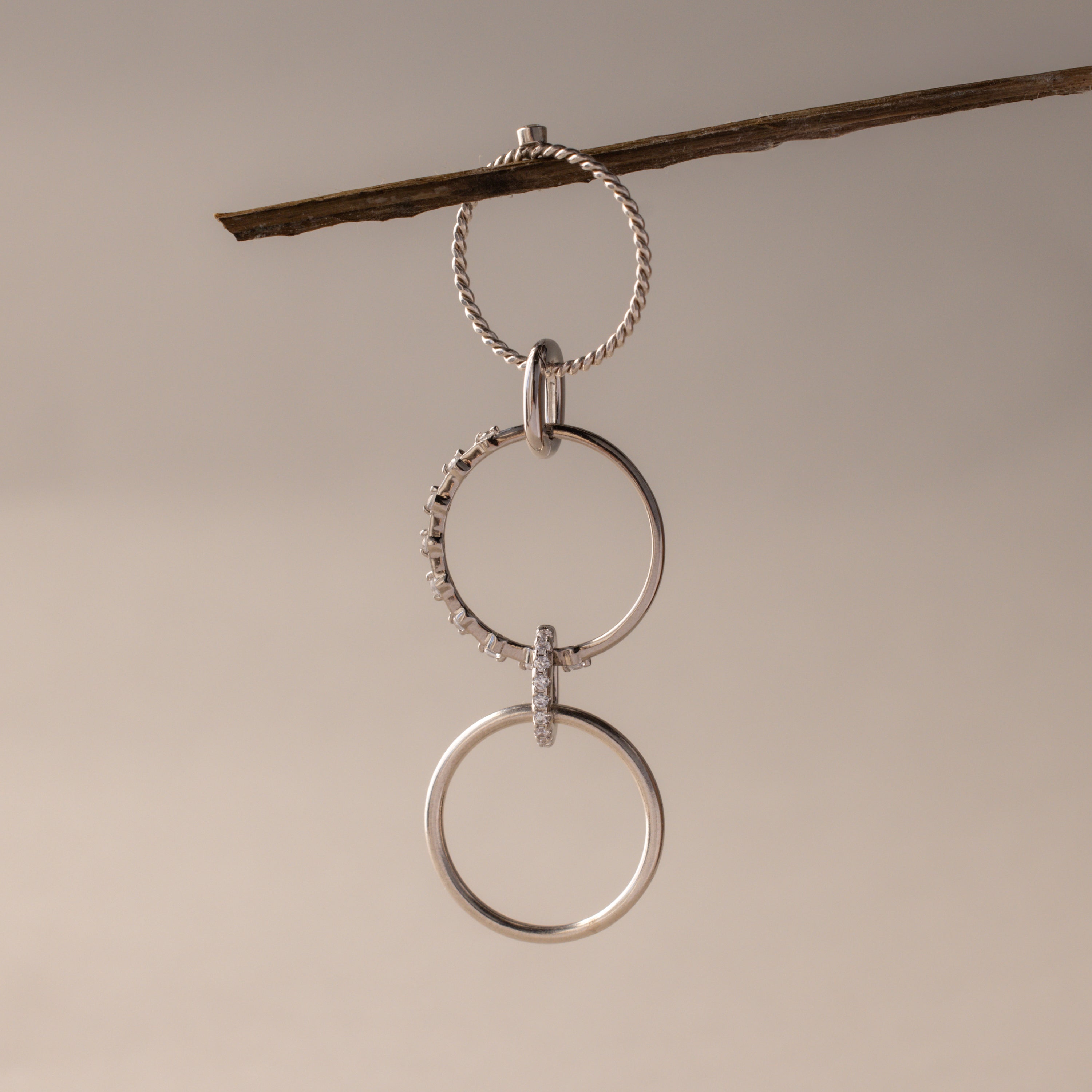 Three silver rings, including the Multipurpose Pave Ring Lock Set in White Gold, hang linked from a twig against a neutral background—highlighting versatile jewelry customization.