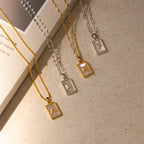 Four Pearl Initial Tag Necklaces, each with a rectangular pendant showing the letters A, E, B, and P, rest on a beige surface near a book—a personalized gift with customizable chain length.