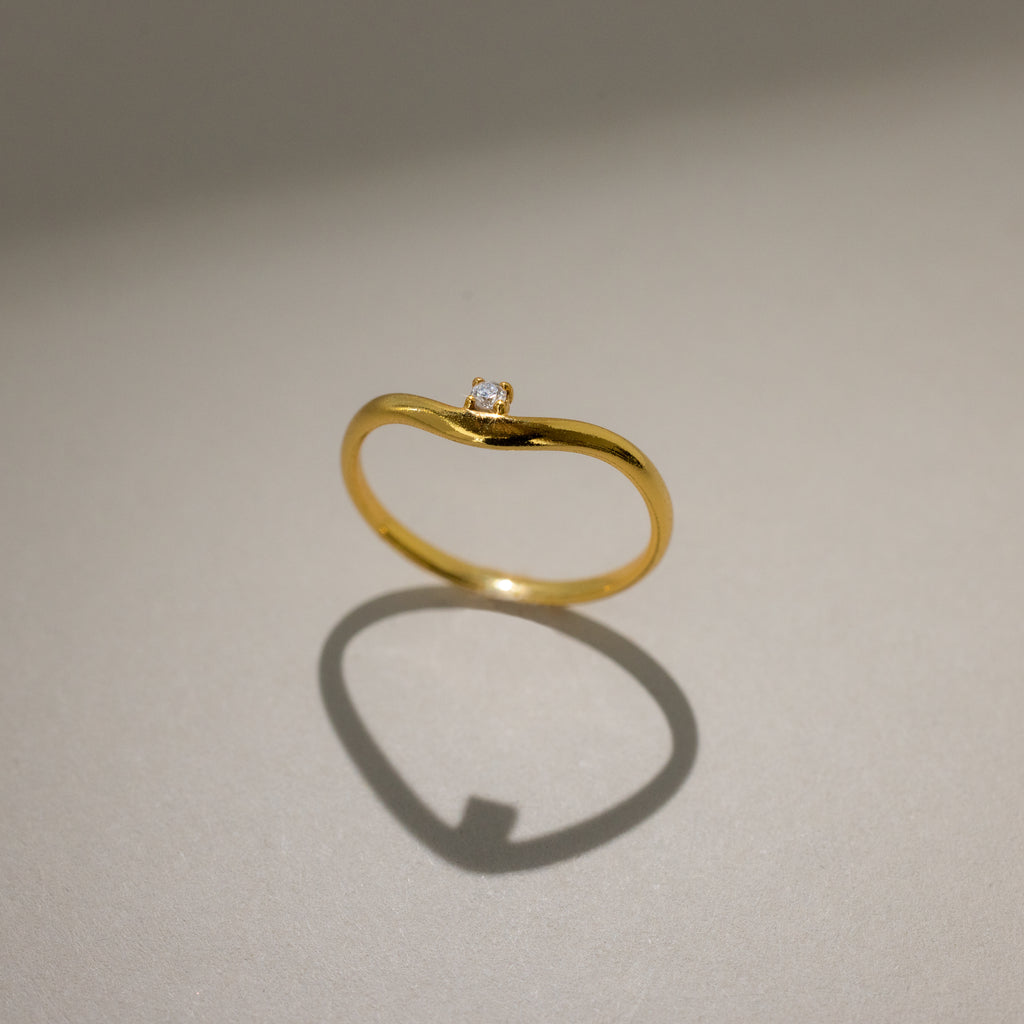 The Diamond Drop Curve Ring, a delicate gold stacking ring with a small diamond, casts a heart-shaped shadow on a light surface.