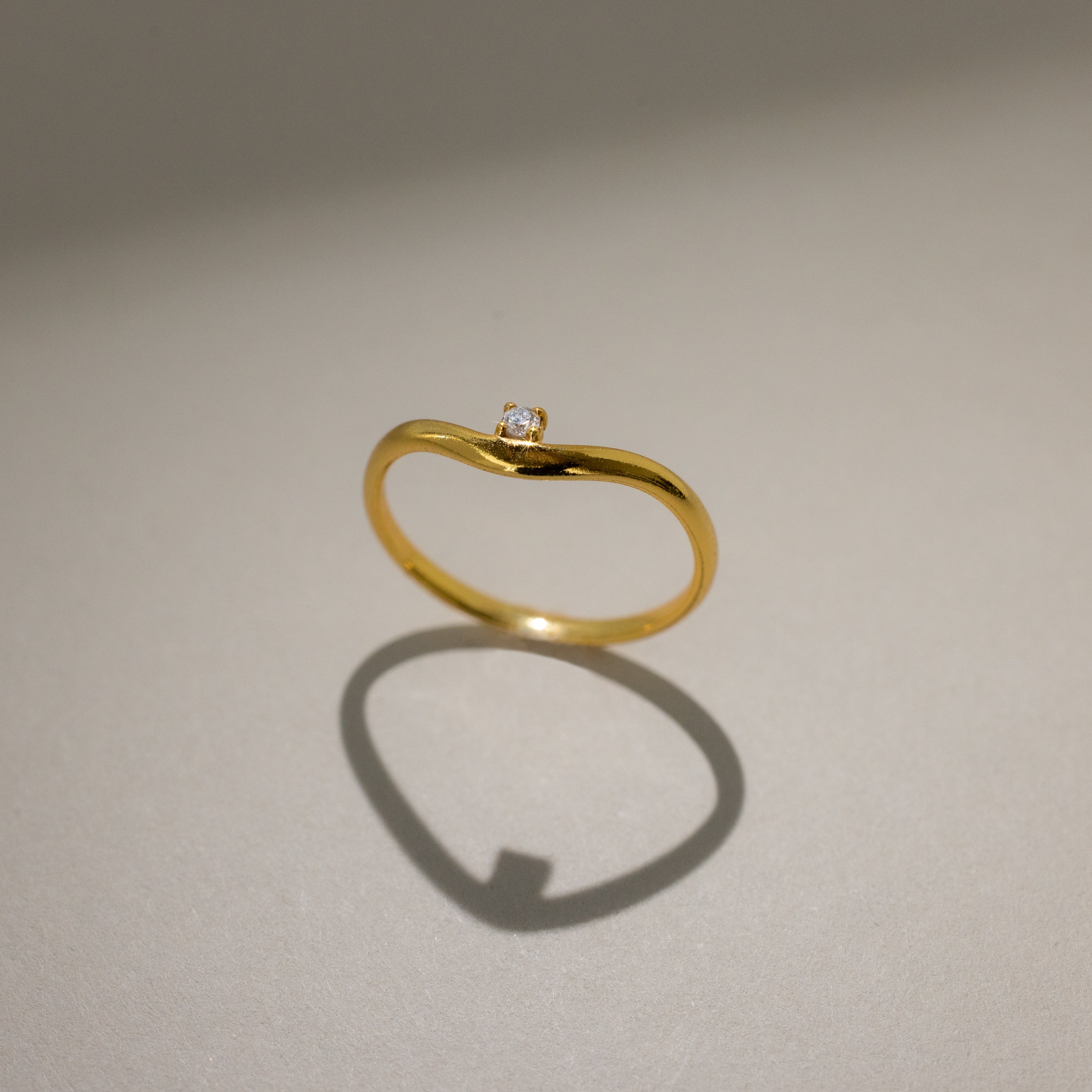 The Diamond Drop Curve Ring, a delicate gold stacking ring with a small diamond, casts a heart-shaped shadow on a light surface.