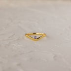The Alexandrite Drop Curve Ring, featuring a delicate gold band with an alexandrite stone at the center, is displayed on a textured white surface.